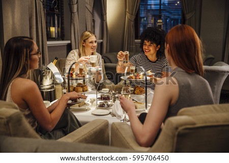 Four women are sitting together enjoying afternoon tea. They are talking and eating and they have champagne and tea. 