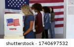 Four women of various demographics, young blonde woman in front, filling in ballots and casting votes in booths at polling station, US flag on wall at back. Focus on booth signage