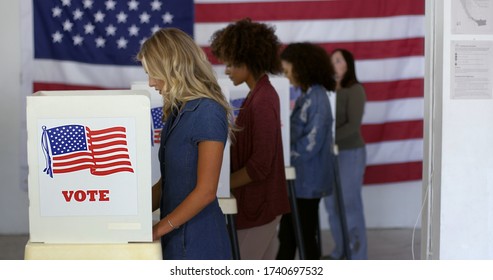 Four women of various demographics, young blonde woman in front, filling in ballots and casting votes in booths at polling station, US flag on wall at back. Focus on booth signage
