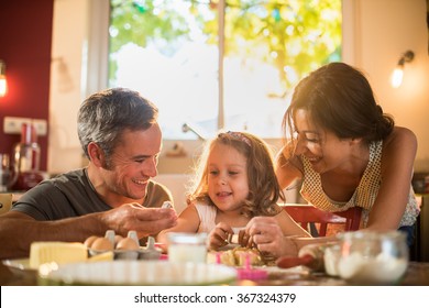 A Four Years Old Blonde Girl Is Cooking Small Cakes With Her Parents In A Luminous Kitchen. They Are Sitting At A Wooden Table, The Dad And The Little Girl Are Holding  Funny Smalls Baking Molds