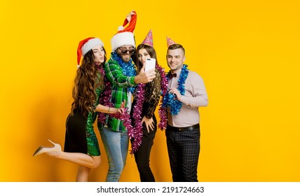 Four Young Adults 20s Wearing Party Hats And Santa Claus Caps And Tinsel Filming Or Taking A Selfie For Social Media On A Yellow Isolated Background With Copy Space