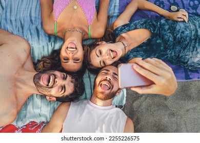 Four young adults enjoying a day at the beach, lying on towels and taking a group selfie. Laughing and making silly faces, they capture the joy and memories of a perfect summer day spent with friends - Powered by Shutterstock