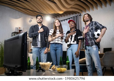 Four young friends keeping hands on hearts singing hymn before start watching football game on TV. Multiracial soccer fans gathering together at home to support favorite team during world cup.