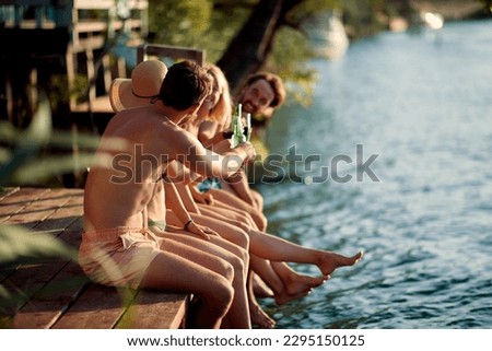 Four young friends on vacation together. Sitting on wooden jetty by water cheering with drinks. Holiday, togetherness, fun, lifestyle concept.