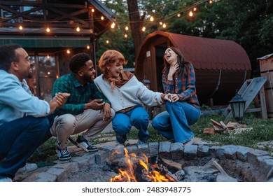 Four young people, two white women and two black men having fun and laughing while squatting next to a burning fire pit in a backyard. - Powered by Shutterstock