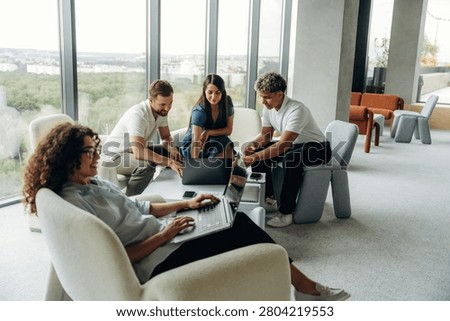 Four young professionals engage in a collaborative meeting in a contemporary office, with one woman focused on her laptop while others share ideas