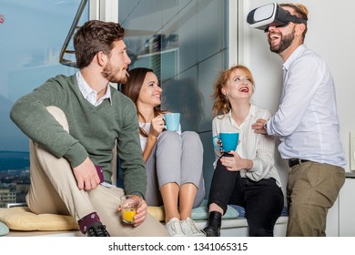 Four Young Professionals Have A Break And Making Fun Jokes With Vr Goggles. They Drink Coffee And Tea And Lead One Colleage. Casual Friday After Work Concept.