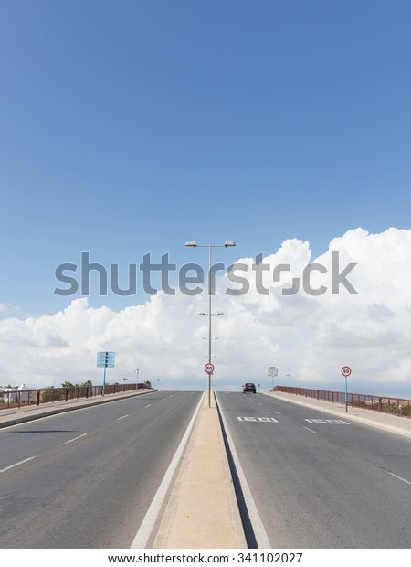 four-lane asphalt road with lamp posts goes beyond\
the horizon with blue sky and the black car rides and road signs\
restricting speed