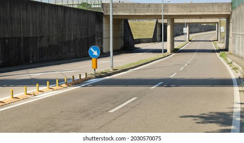 A four-lane road, with barrier on the middle, passes under a  gray concrete underpass and alongside a wall of the same color. Concrete sidewalk and traffic signs. Background for copy space