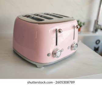 four-slot toaster in pastel pink with chrome accents and a retro design. It has two control levers and two dials to adjust the toasting levels. The toaster is placed on a white countertop