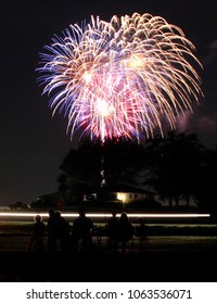 Fourth of July fireworks erupt in countryside while family watches in foreground. 