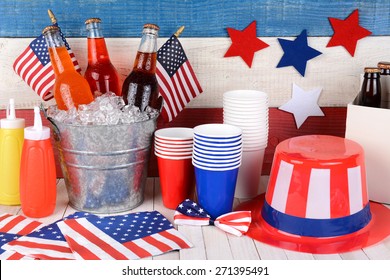 Fourth Of July Still Life. A Picnic Table With A Bucket Of Soda, Flags, Uncle Sam Hat, 6 Pack, Ketchup And Mustard With A Red, White And Blue Wall Behind.