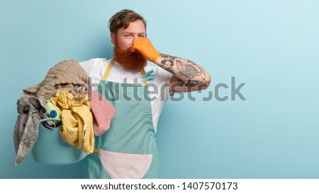 Foxy bearded man covers nose, holds pile of dity laundry in basin, smells unpleasant stinch, has tattoo on arm, has to do washing, isolated over blue background, free space aside for your advert