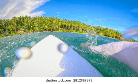 FPV, POV: View from a surfboard while paddling through turquoise ocean toward tropical sandy beach covered with tall palm trees. Splashes and flying water droplets as surfer moves in the lineup.