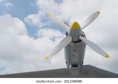 A fragment of an airplane wing with a four-bladed white aircraft propeller with yellow tips mounted on it on a cylindrical engine turbine against a cloudy sky.