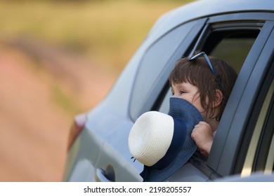 A Fragment Of A Car With A Child In The Rear Side Window. A Smiling Girl Covers Her Face With A Hat. Selective Focus.                               