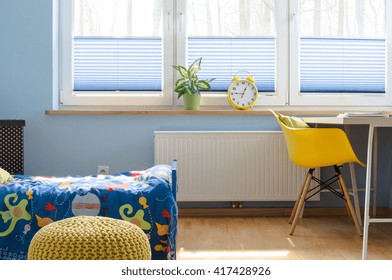 Fragment of a child room with large blinded window, radiator underneath, covered bed and desk with chair beside it