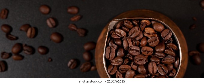 Fragment of a crucible filled with coffee beans, black tabletop with scattered coffee beans in the background.