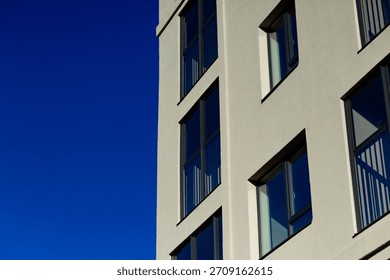 A fragment of the facade of a modern building with rectangular windows against a bright blue sky. Clean lines, light-colored walls, and a laconic architectural style.