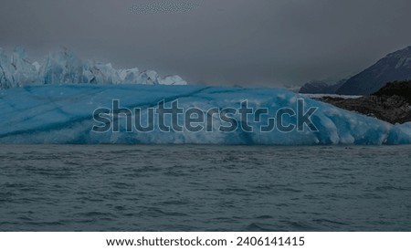 A fragment of a glacier. A wall of blue ice with sharp peaks and cracks and a huge melting iceberg in a glacial turquoise lake. Mountains against a cloudy sky. Fog. Perito Moreno glacier. Argentina. 