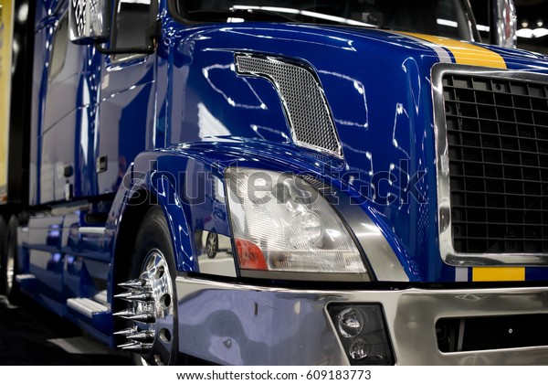A fragment of a large commercial semi truck with\
modern design of headlights and a grille, with chrome-plated rims\
and custom bolt design, a shining bumper and a blue lacquered paint\
finish with glare