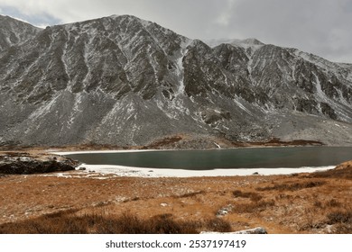 A fragment of a large lake with ice near the shores lying at the foot of a high cliff sprinkled with the first snow on an autumn cloudy day. Lake Kok-Kol, Altai, Siberia, Russia. - Powered by Shutterstock