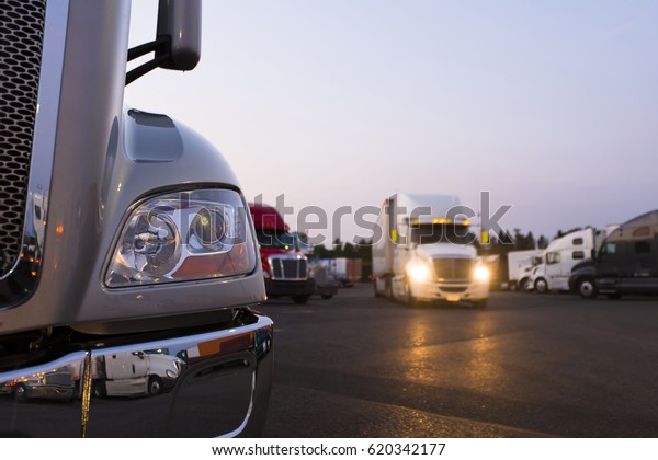 Fragment\
of the modern parts of the truck on a night truck stop in the\
foreground to the background of semi trucks at the parking places\
and a moving semi truck with its headlights\
on.