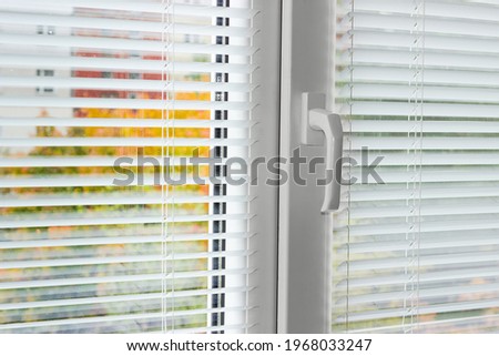 Fragment of the modern plastic window with white horizontal blinds and mosquito net, blurred view of trees across the slats from the inside
