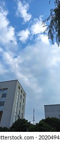 A Fragment Of A Part Of A Television Tower Between Two Buildings And Blue Sky With White Clouds In The City Street In Summer.