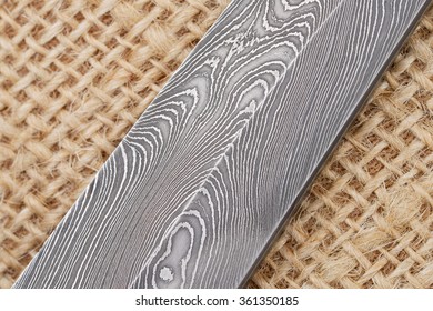 Fragment of the traditional handmade Finnish knife blade with the abstract wave pattern of damascus steel over an old sack background. 