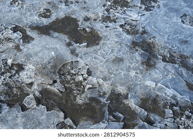 Fragmented Ice Fields With Overlapping Translucent Sheets And Exposed Dark Soil Creating Mosaic Of Cracked Frozen Surface In Cold Outdoor Environment Under Diffused Light.