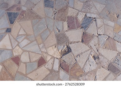 Fragmented stone tiles create an irregular mosaic pattern. Earthy tones include beige, gray, and soft reds. Irregular shapes add natural texture to the surface. The design feels rustic and organic