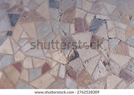 Fragmented stone tiles create an irregular mosaic pattern. Earthy tones include beige, gray, and soft reds. Irregular shapes add natural texture to the surface. The design feels rustic and organic