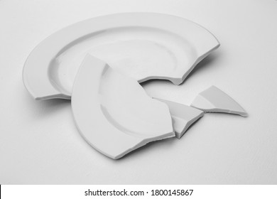 Fragments Of A Broken Plate On A Light Background, Black And White Photo. The Concept Of Breaking Up Relations, Divorce, Destruction