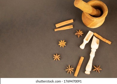 Fragrant Spices And Kitchen Utensils. On A Brown Background Are Spread Cinnamon Sticks And Star Anise. In The Corner There Is A Mortar For Spices Made Of Honey Oak And Two Spoons Made Of Light Wood.