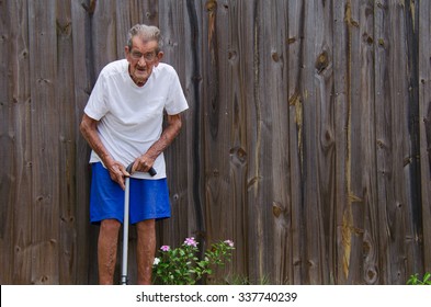 A Frail One Hundred Year Old Centenarian Senior Citizen Man With A Cane Standing By A Wooden Fence. Ample Room For Text In Negative Space Over The Fence.