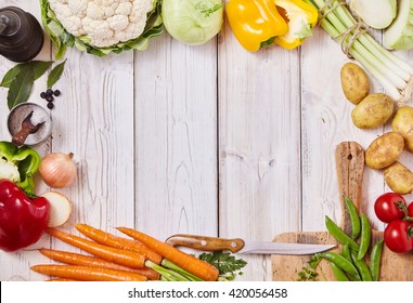 Frame Of Assorted Healthy Fresh Farm Vegetables, A Cutting Board, Knife, Salt And Pepper Mill On A White Wooden Background With Central Copy Space, Overhead View