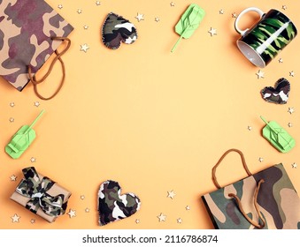 Frame of camouflage hearts, gifts, bag, mug and origami paper tanks on yellow background. Top down composition with cope space. Defender of the Fatherland Day, February 23. - Shutterstock ID 2116786874