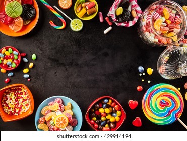 Frame of colorful bright assorted candy in bowls and jars, candy canes and rainbow colored spiral lollipops on black with scattered candy hearts and jellybeans around a central copy space on slate - Powered by Shutterstock