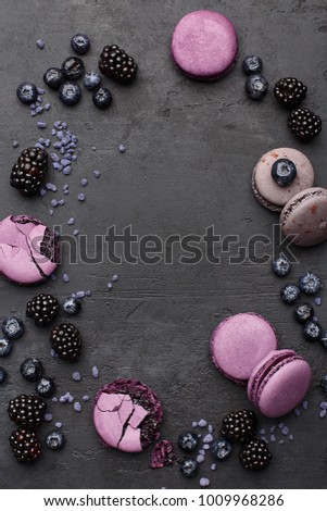 Frame composition made of violet and lilac macaroons, fresh blackberries and blueberries on black concrete background with copy space. Flat lay, food background. Traditional French dessert