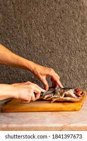 In The Frame Of Female Hands With A Carving Knife.  Frozen Fish Silver Hake On A Wooden Board.  Background Gray, Structural.