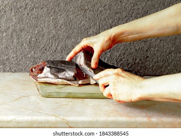 In The Frame Of Female Hands With A Carving Knife.  Frozen Fish Silver Hake On A Wooden Board.  Background Gray, Structural.  Beautiful Shot.