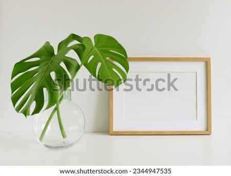 Frame mockup, glas vase with green monstera leaves onwhite table near white wall. Photo frame, poster template . Copy space.