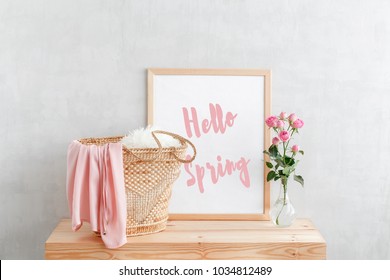 Frame With Text HELLO SPRING, Woven Straw Bag And Vase With Pink Rose Flowers On A Wooden Table On A Background Of Light Gray Walls. Home Interior Decor.