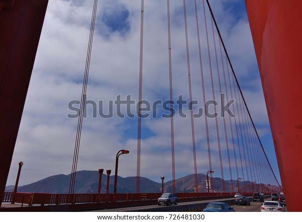 Framed on left & right by the base of a Golden Gate
Bridge tower this is a view on the bridge looking through
suspension cables at Marin Headlands and blue sky with white
clouds. Some traffic seen.
