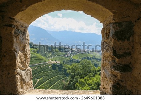 Framed view through Tyrol Castle’s window shows Dorf Tirol’s terraced vineyards and church under sunny skies, with hazy Alpine foothills beyond. South Tyrol, Italy.