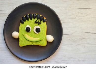 Frankenstein Avocado Toast Made From Toast,mashed Avocado,mozzarella Balls And Black Olives On Black Plate With White Wood Table Background.Art Food Idea For Kids Party.Top View.Copy Space