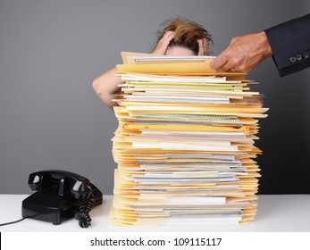 A frazzled female office worker grabs her head in frustration as her boss piles more work on her desk. Horizontal format over a gray background.