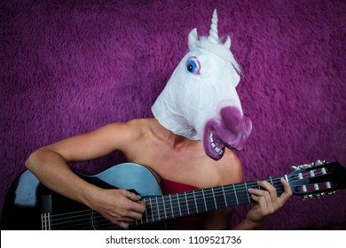 Freaky young woman in comical mask playing the guitar on the purple background. Portrait of unusual lady in red dress with musical instrument. Musical performance.
