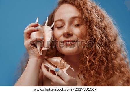 Freckled redhead plump plus size woman holding seashells isolated on blue background. The concept of face and body skin care, body positive and cosmetology.
